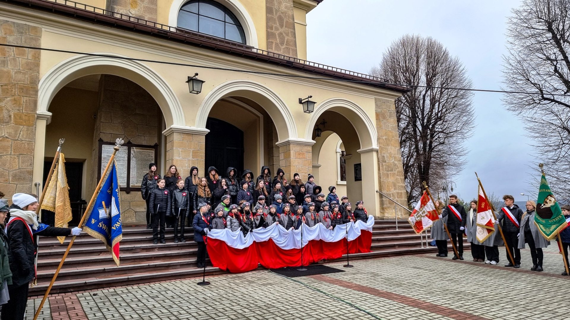 Grupa młodzieży stoi na placu kościelnym z rozciągniętą flagą RP, po lewej i prawej stronie stoją delegacje pocztów sztandarowych.