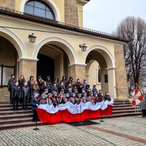 Grupa młodzieży stoi na placu kościelnym z rozciągniętą flagą RP, po lewej i prawej stronie stoją delegacje pocztów sztandarowych.